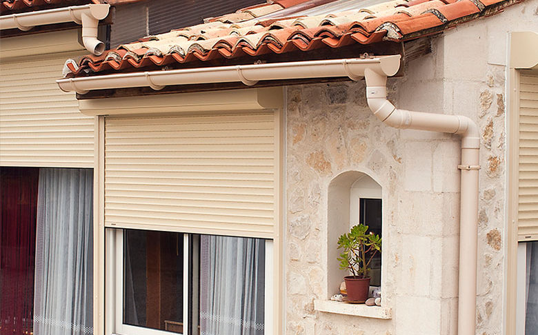 Volets roulants beiges sur une maison en pierre, sous la gouttière